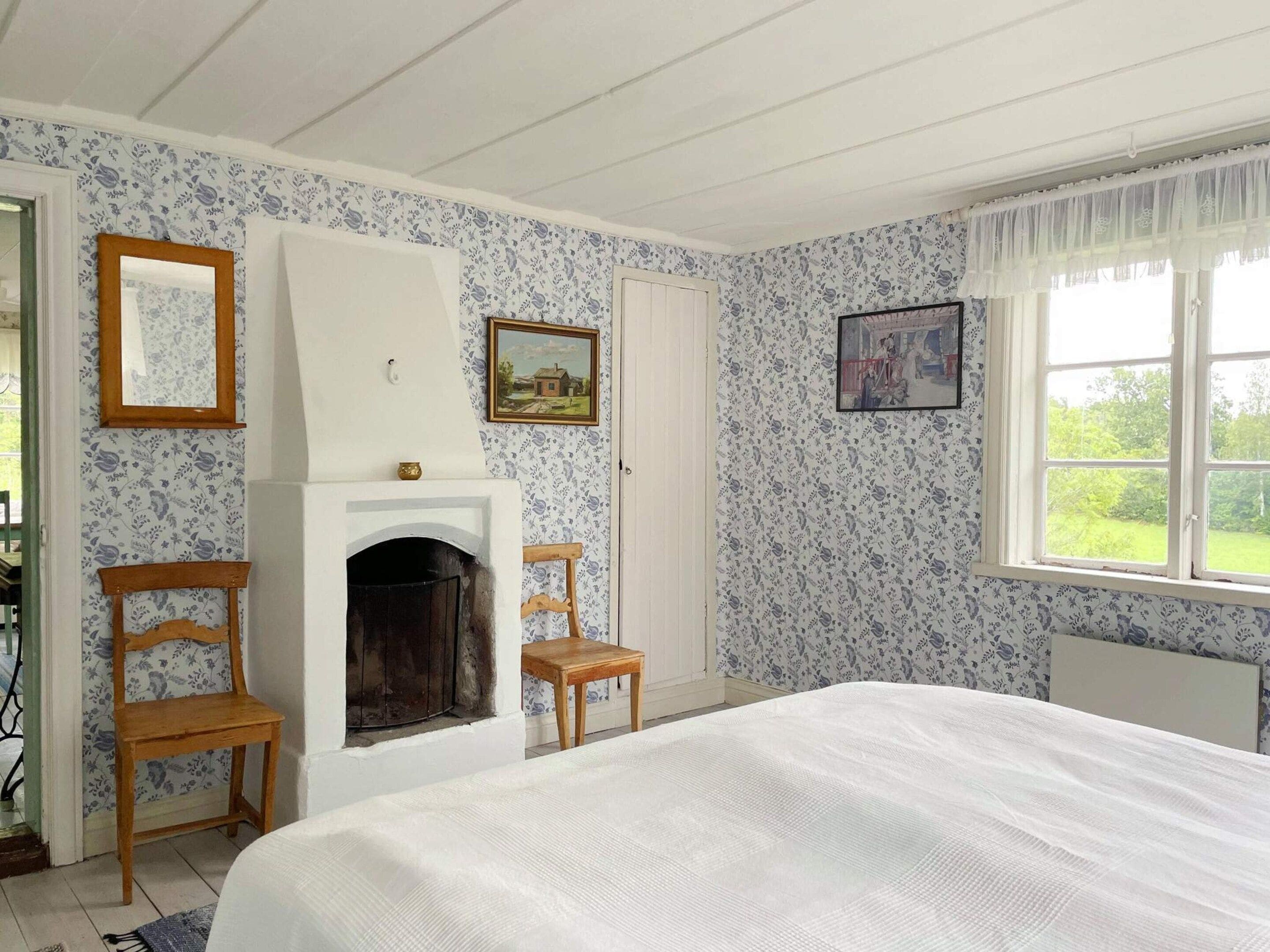 A bedroom with a fireplace, two wooden chairs, a bed, and floral wallpaper.
