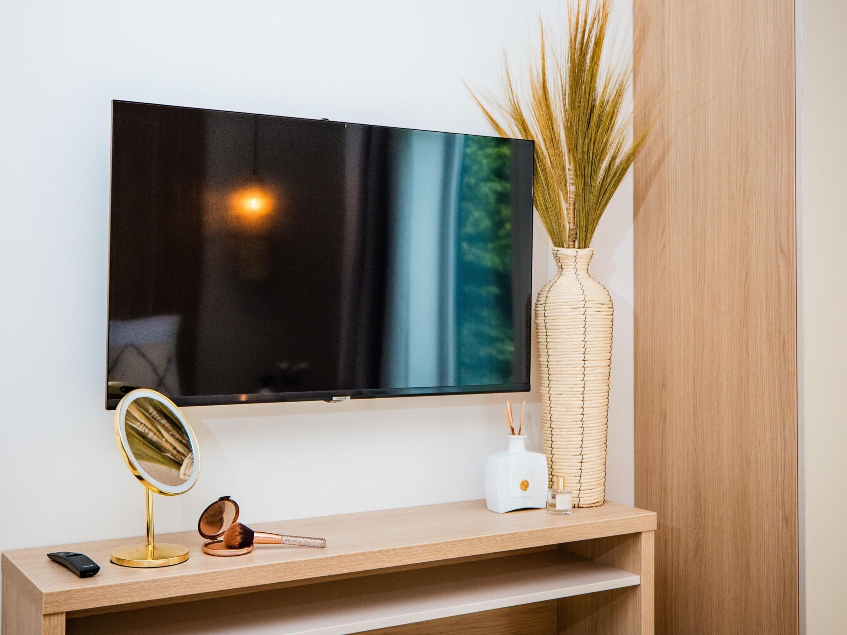 Ein Flachbildfernseher an der Wand, ein runder Spiegel, ein Make-up-Pinsel, ein Smartphone, eine weiße Vase mit Schilf und ein weißer Diffusor auf einer Holzkonsole.