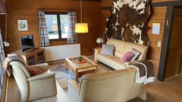 Ein gemütliches Wohnzimmer mit einer Couch, zwei Sesseln, einem Couchtisch und einem Fernseher auf einem Holztisch. An der Wand hängt ein großes Fell, und es gibt ein Fenster mit Vorhängen.