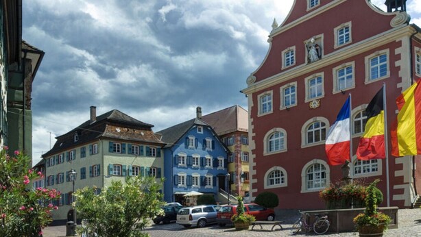 Une place pavée, bordée de bâtiments historiques aux couleurs vives, où flottent des drapeaux, et où l’on aperçoit un vélo.