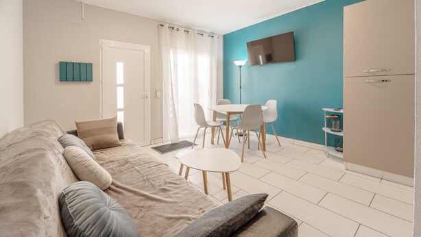 A modern living room with a beige sofa, a dining area with a white table and chairs, a teal accent wall, and a built-in TV. - WELCS CASA ADOSADA 144 EMP con piscina comunitaria (Rosas / Roses)