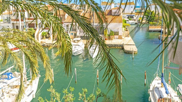 A marina with boats docked, surrounded by buildings with red-tiled roofs and palm trees. - WELCS APARTAMENTO 145 EMP con vistas al canal (Empuriabrava)