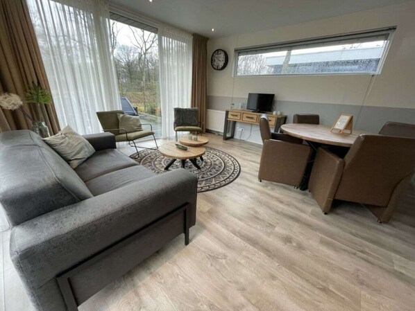 A modern living room with a grey sofa, a round coffee table, a TV, and a dining area with a table and chairs. - Pavilion 4 People (Zorgvlied)