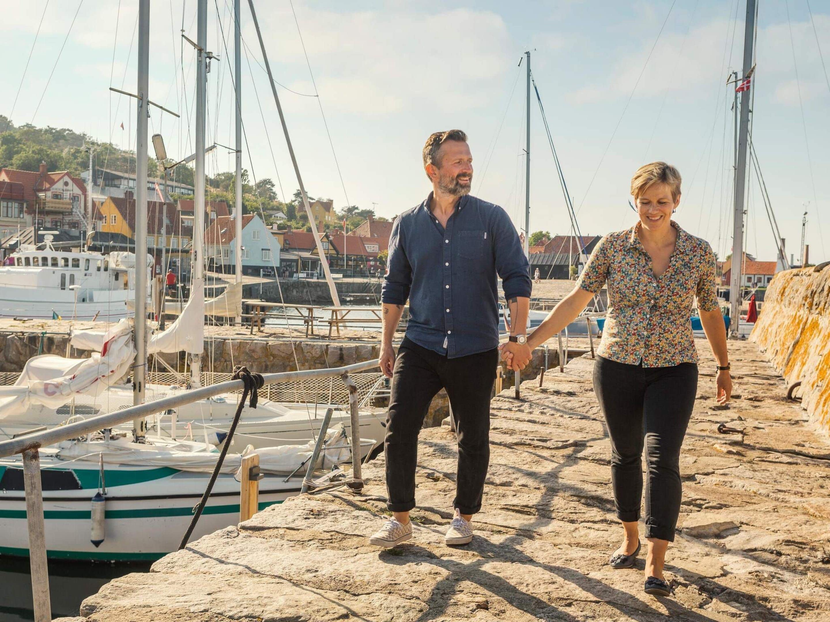 A couple walking on a stone pier with boats and buildings in the background.