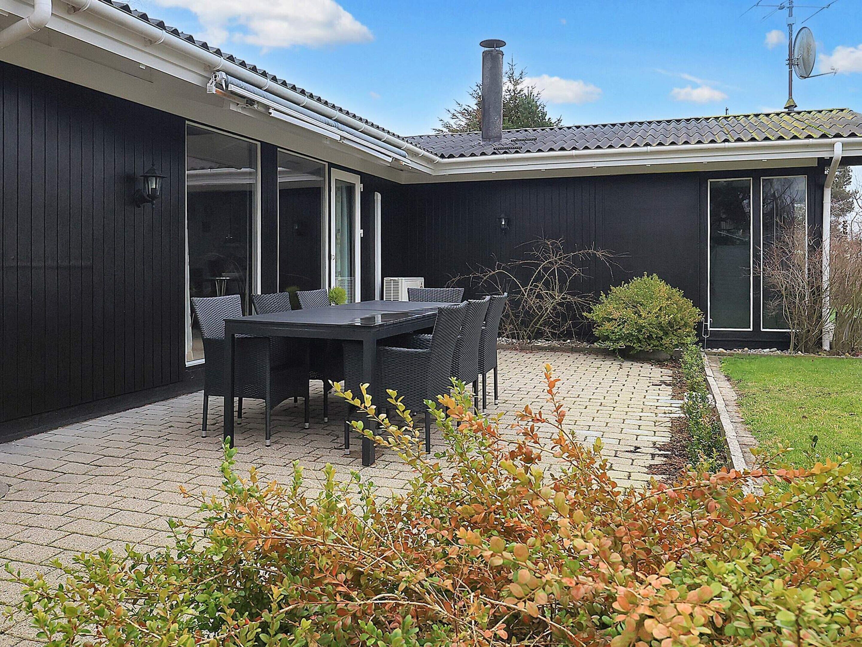 A modern outdoor dining area with a black table and chairs, surrounded by a brick patio and greenery.