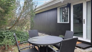 A patio with a round table and four chairs, a sliding glass door, and a dark-colored building facade. - 4 Star Holiday Home in Vaeggerlose (Vaeggerlose)