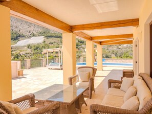 A covered outdoor seating area with wicker furniture, a glass table, and a view of a pool and hills. - Belvilla by OYO Villa Nancy (Callosa d' En Sarria)