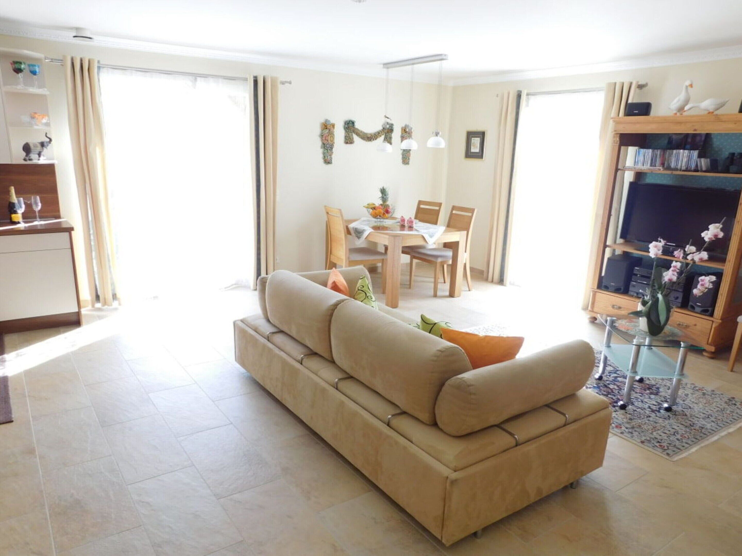 A living room with a sofa, a dining table with chairs, a TV stand with speakers, and a bookshelf.
