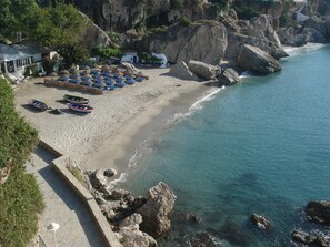 Una platja amb filera de gandules blaves i blanques, un edifici petit i penya-segats rocosos.