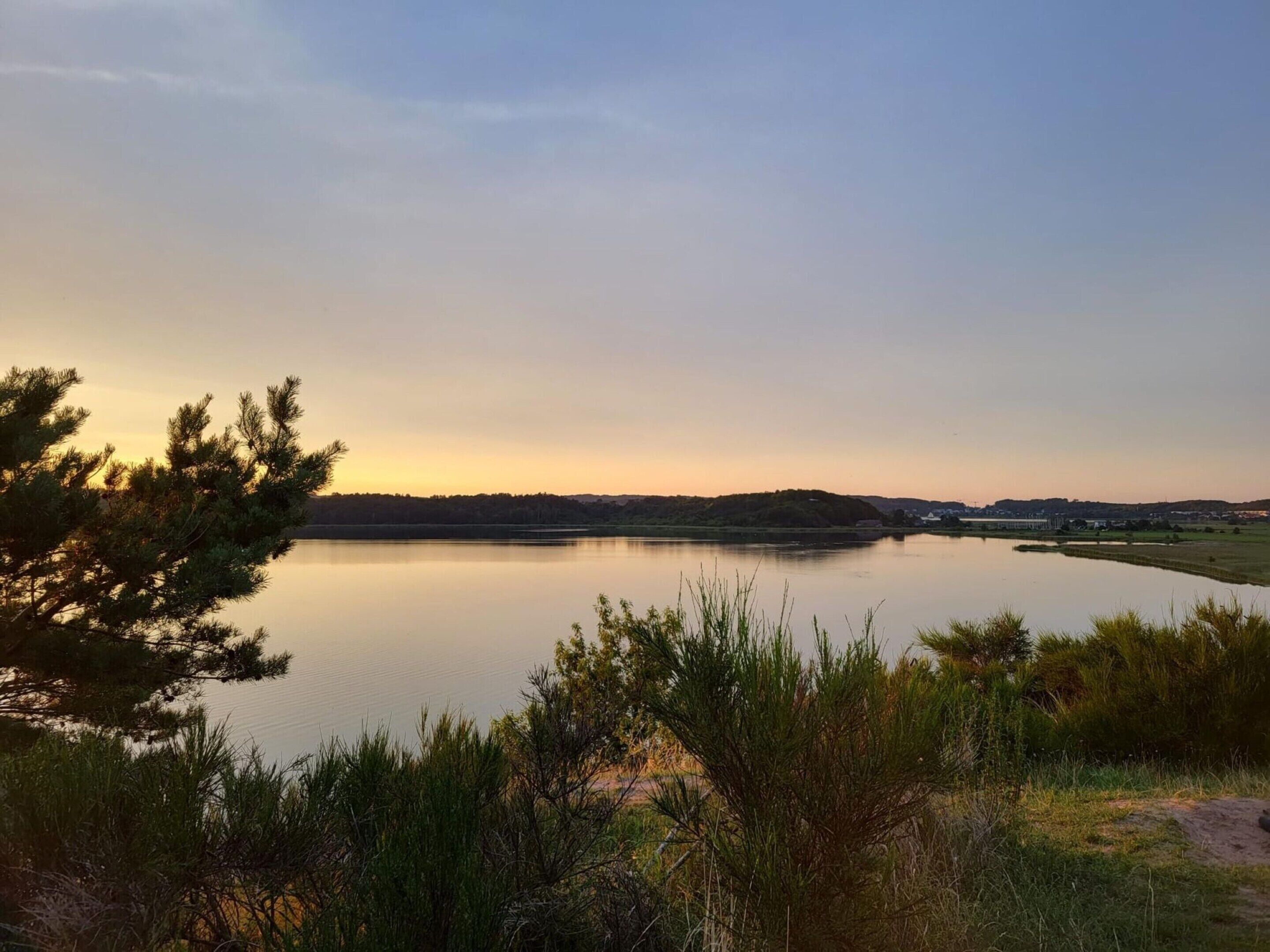 A serene lakeside view with a clear sky, calm water, and surrounding vegetation.