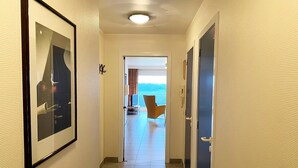 En hotellkorridor med en inramad tavla på vänster sida, en dörr på höger sida och en utsikt över en balkong med en stol och ett bord.