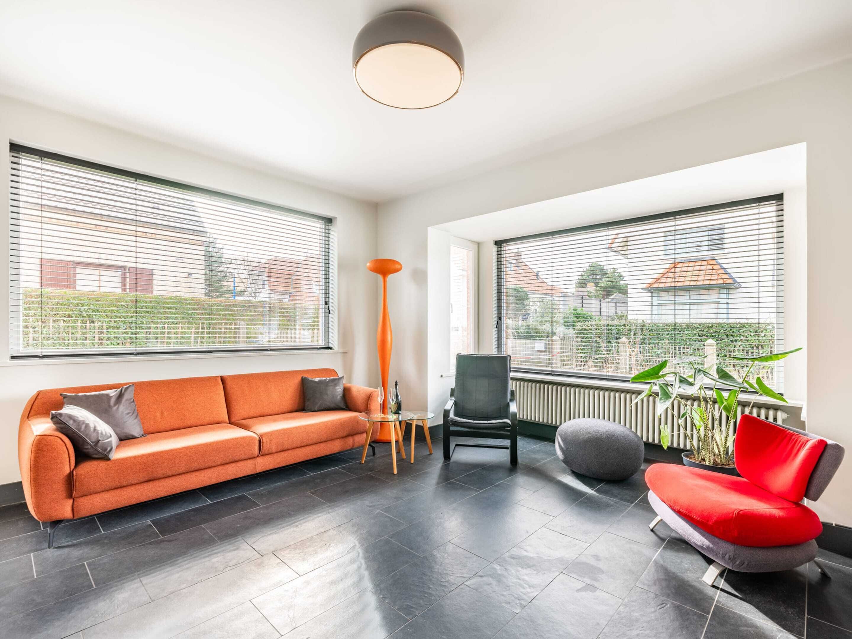 A modern living room with a sofa, chair, and a red armchair. There are large windows with blinds, a radiator, and a floor lamp.