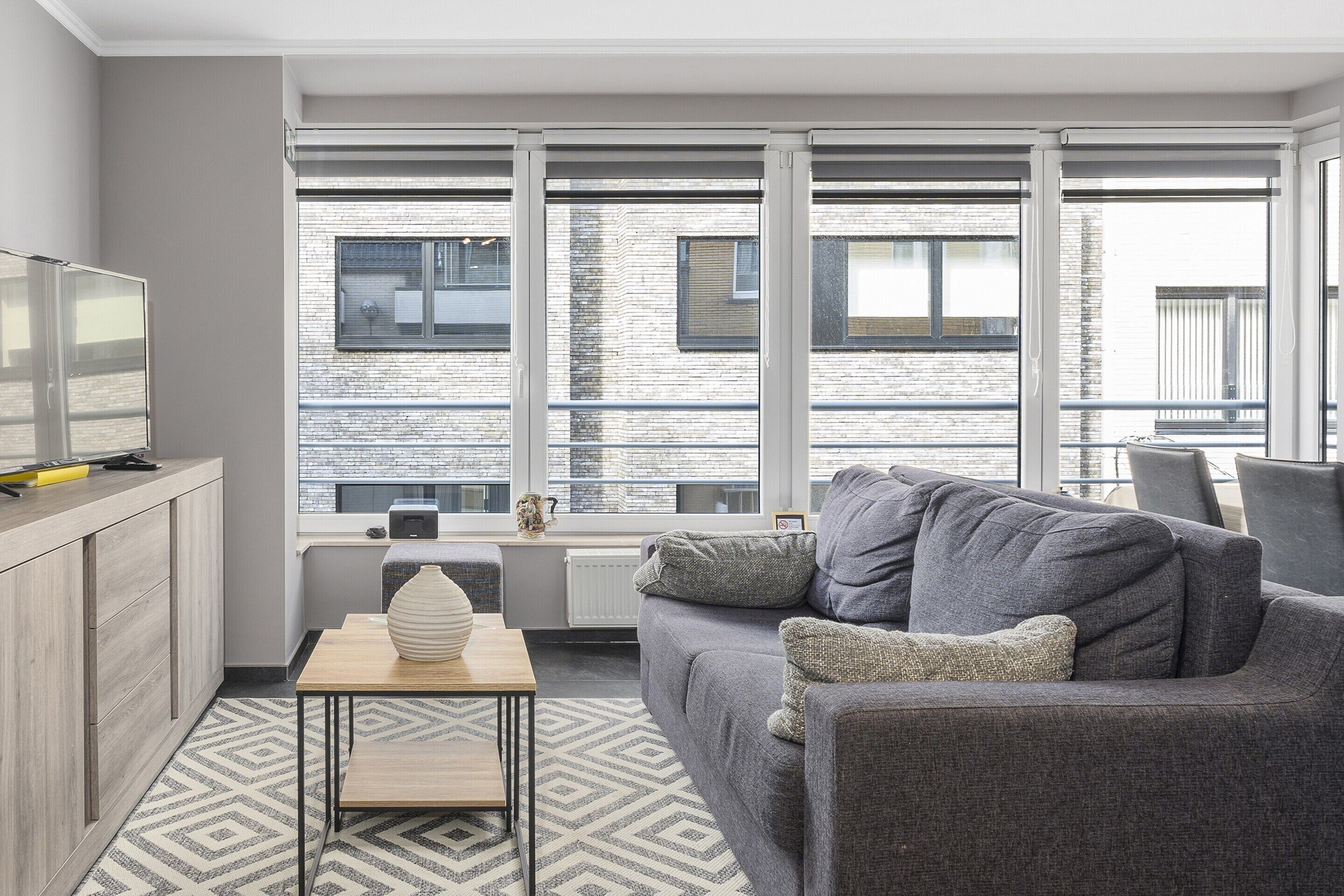 A modern living room with a grey sofa, a small table with a vase, and a view of a brick building through large windows.
