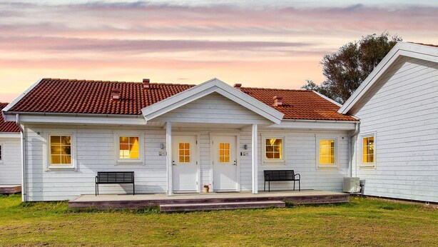 A two-story white house with a red-tiled roof, numbered 15 and 16, featuring a front porch with benches and a small garden area.