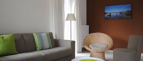 A modern living room with a sofa, a wicker chair, a round ottoman, and a painting on the wall.