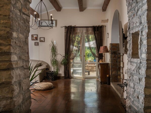 Quarto com paredes de pedra, teto de madeira, um lustre, sofá, uma luminária e vista para área externa através de uma porta de vidro.