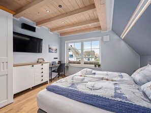 Une chambre à coucher comprenant un lit, un bureau, une chaise, un téléviseur et une fenêtre donnant sur des maisons.