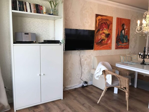 A room with a white cabinet, a flat-screen TV mounted on the wall, a wicker chair, and a table with a candelabra. - Holiday Apartment in Southern Alps (Scharbeutz)