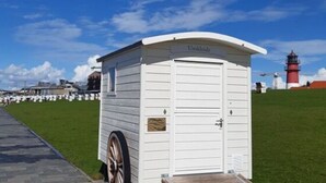 Une maison mobile blanche, équipée d’une rampe d’accès, est située sur un chemin pavé, avec un champ verdoyant et un phare en arrière-plan.