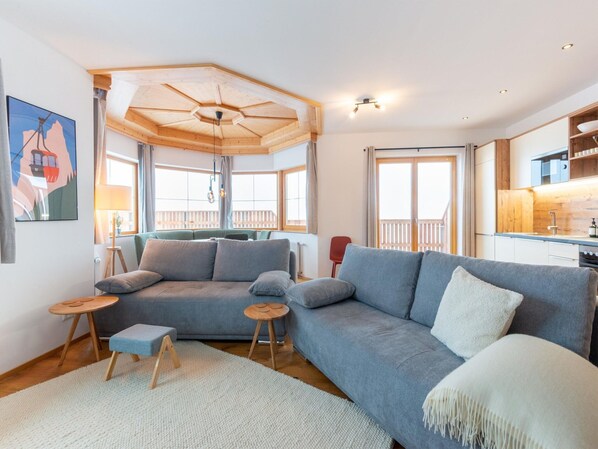 A modern living room with a large gray sectional sofa, a wooden ceiling fan, and a view of the balcony through large windows. - Apartment in Stubai Valley With Mountain View (Neustift Im Stubaital)