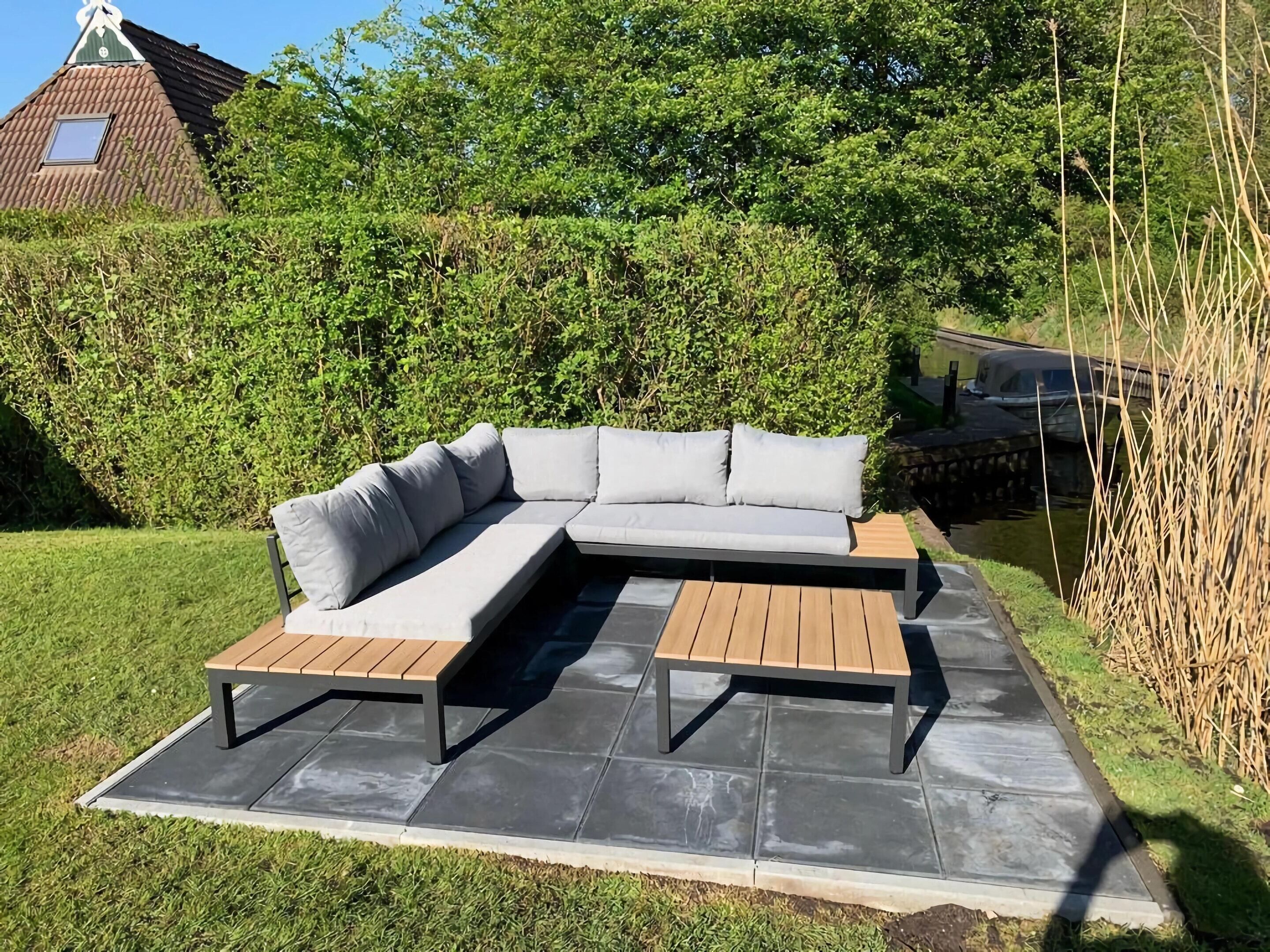 A sectional sofa with cushions on a patio area with a stone floor, surrounded by greenery and a water feature.