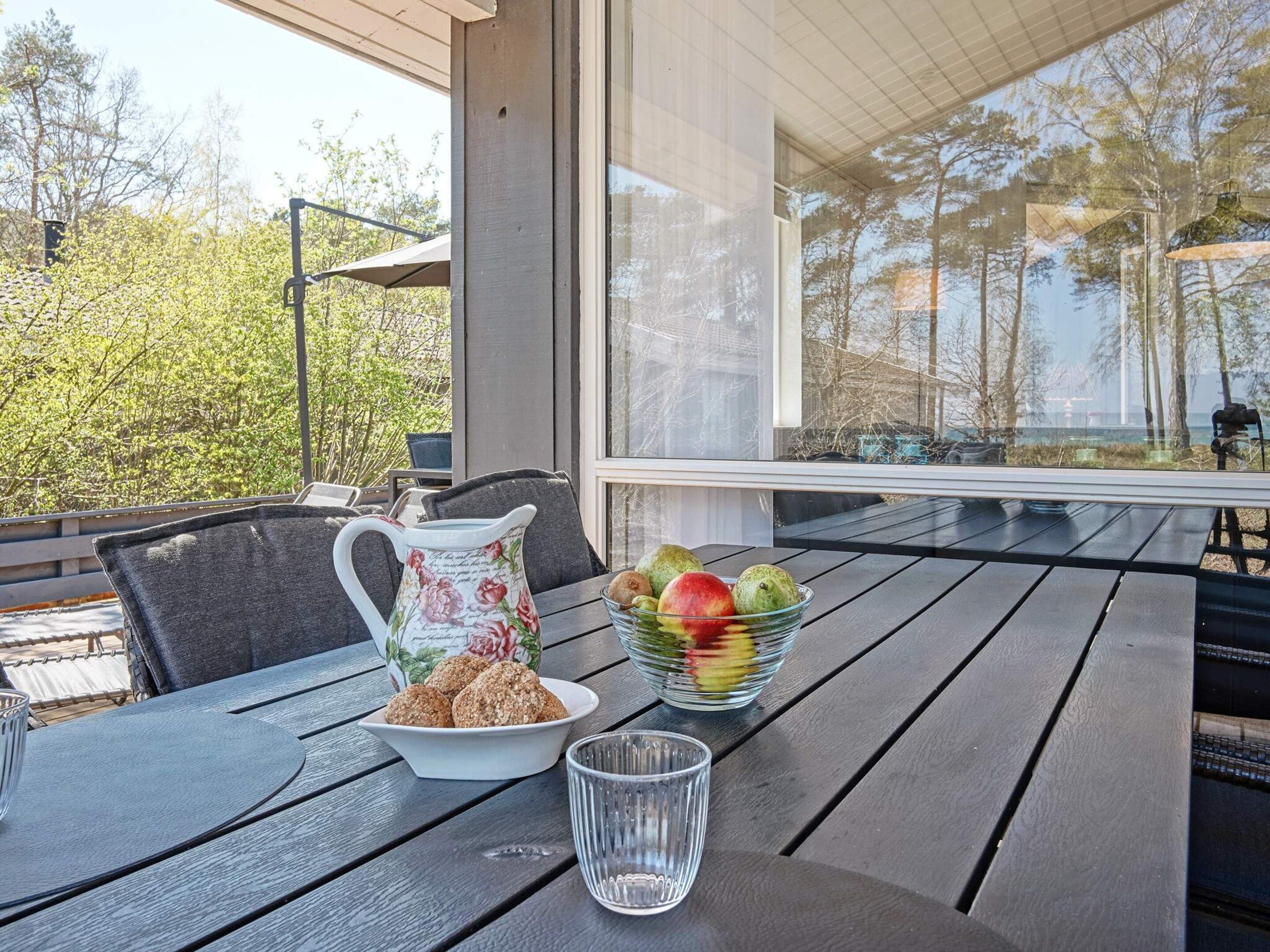 Una mesa con una jarrón, un cuenco de fruta y galletas, al aire libre con vistas a árboles y un edificio en el fondo.