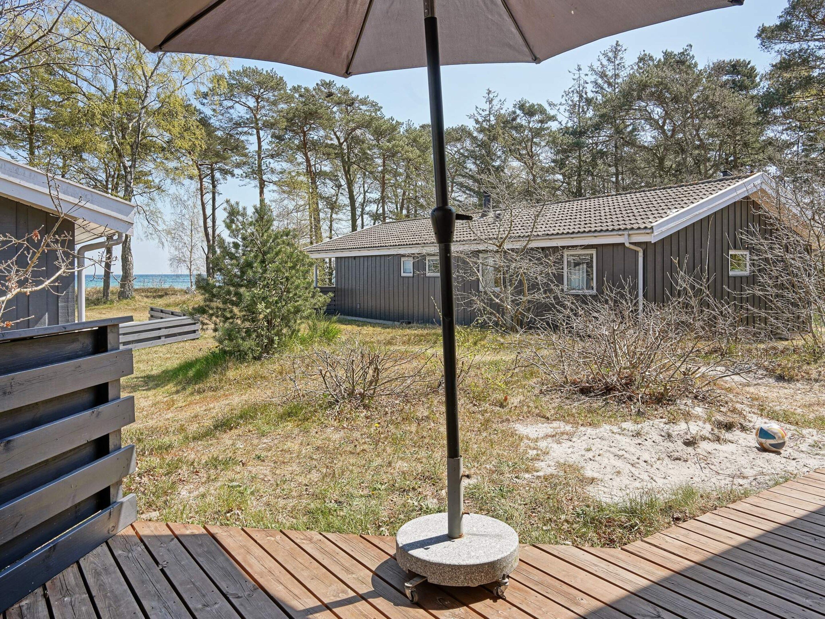 A wooden deck with a stone table and an umbrella, overlooking a beach with cabins and trees.