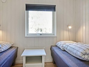 Una habitación con dos camas individuales, una mesita blanca, una ventana con persianas y una lámpara fijada a la pared.