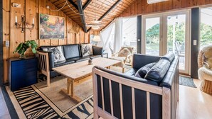 A cozy cabin interior with a wooden ceiling, a sofa set, a dining table, and a large window offering a view of the outdoors.