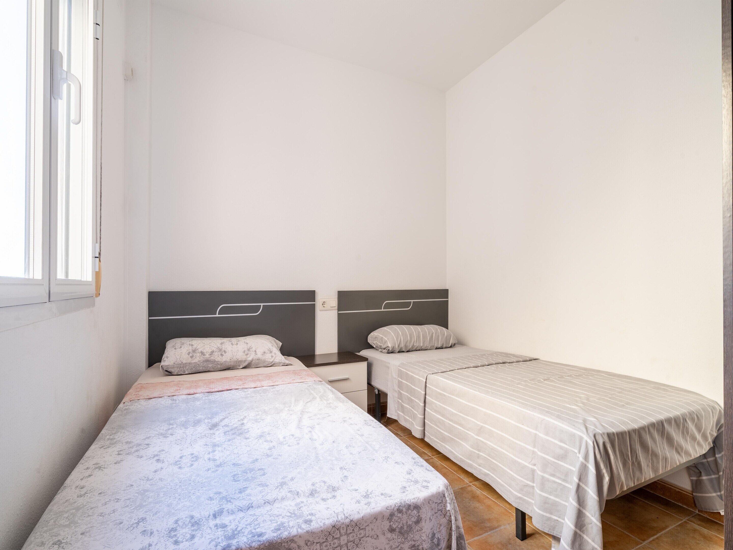 A small, clean room with two single beds, a window, and a plain white wall.