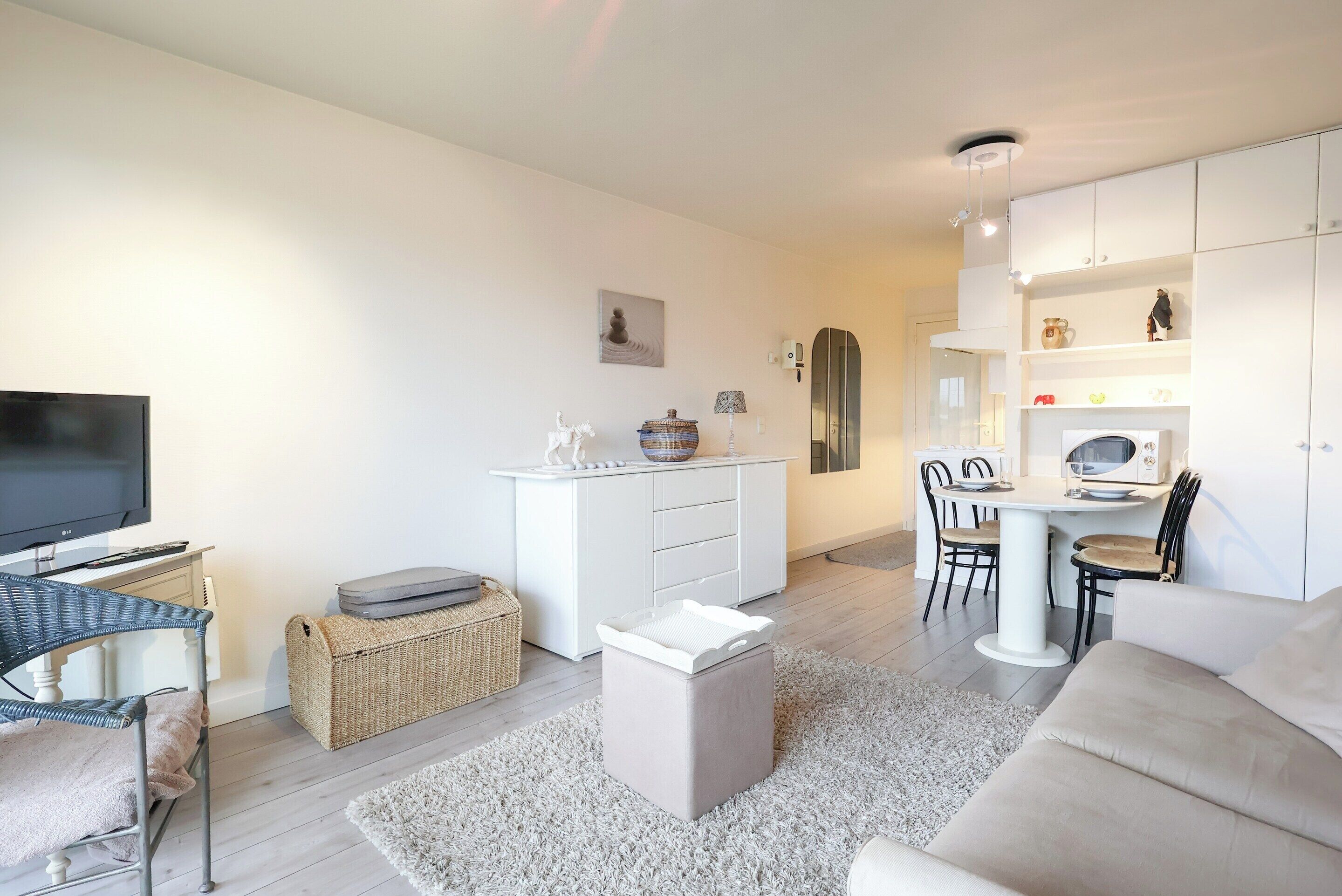 A modern living room with a flat-screen TV, a sofa, a dining area with a table and chairs, and a kitchen area with cabinets and appliances.