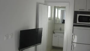 Una habitación pequeña y blanca con televisión de pantalla plana montada en la pared, microondas y lavabo con armarios.