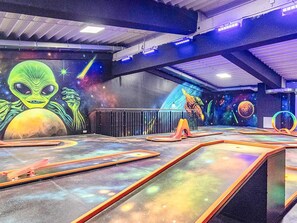 Um espaço interno com um mural de um alienígena e planetas, uma rampa com piso temático espacial e uma estrutura circular.