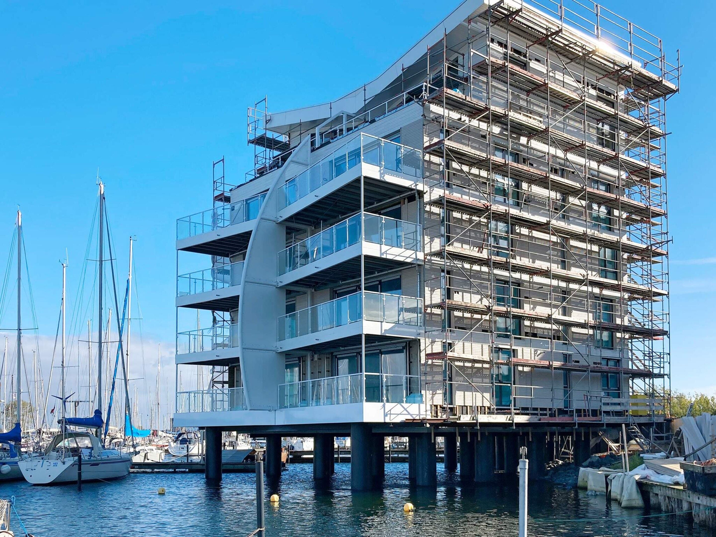 A multi-story building under construction with balconies and large windows, situated over a body of water with boats.