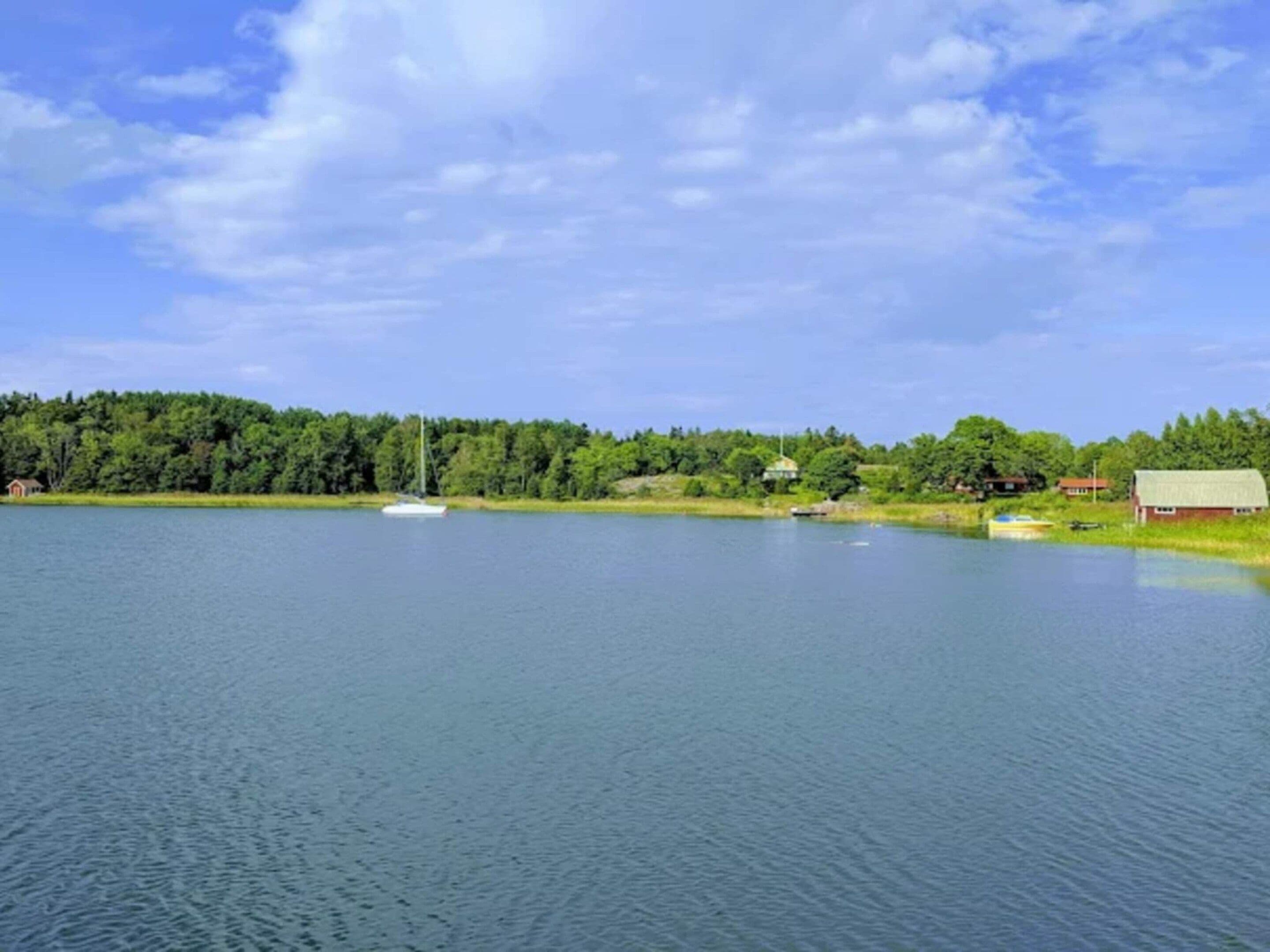 Un lac paisible où flotte un voilier, entouré d’une forêt dense et de quelques bâtiments.