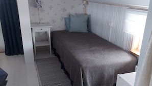 A single bed with a dark-colored bedspread, a bedside table, a small desk, and a window with vertical blinds. - 7 Person Holiday Home in Grisslehamn (Grisslehamn)