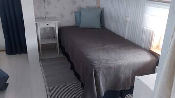 A single bed with a dark-colored bedspread, a bedside table, a small desk, and a window with vertical blinds.