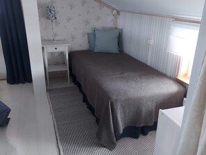 A single bed with a dark-colored bedspread, a bedside table, a small desk, and a window with vertical blinds. - 7 Person Holiday Home in Grisslehamn (Grisslehamn)
