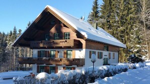 A two-story wooden chalet with a sloped roof, surrounded by snow-covered trees and a clear sky. - Waldhaus Sonnenberg Apartment 1 (Drachselsried)