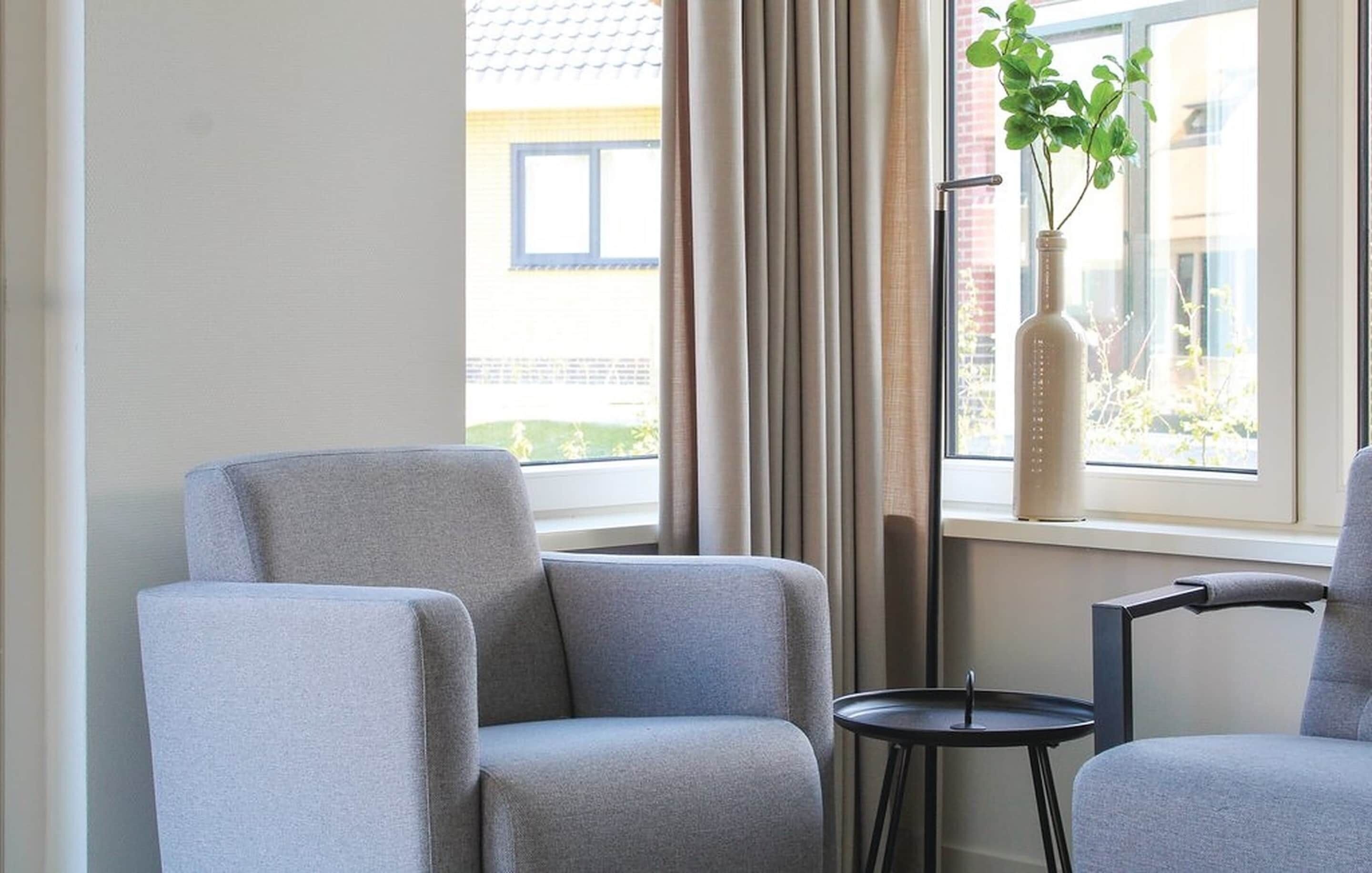 Ein Zimmer mit zwei grauen Sessel, einem kleinen runden Tisch, einer Vase mit Grünpflanzen und einem Fenster mit Vorhängen.