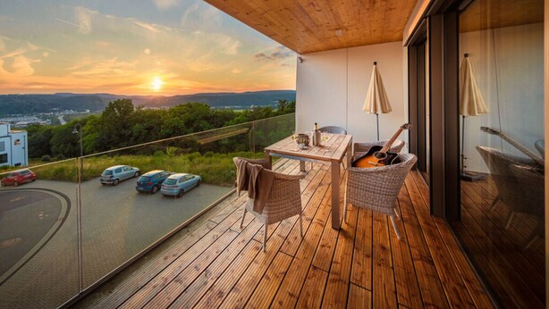 A wooden deck with a table and chairs, a guitar, and a view of the sunset over a landscape. - Mosel Sky Lounge With Loggia & Panoramic View (Kenn)