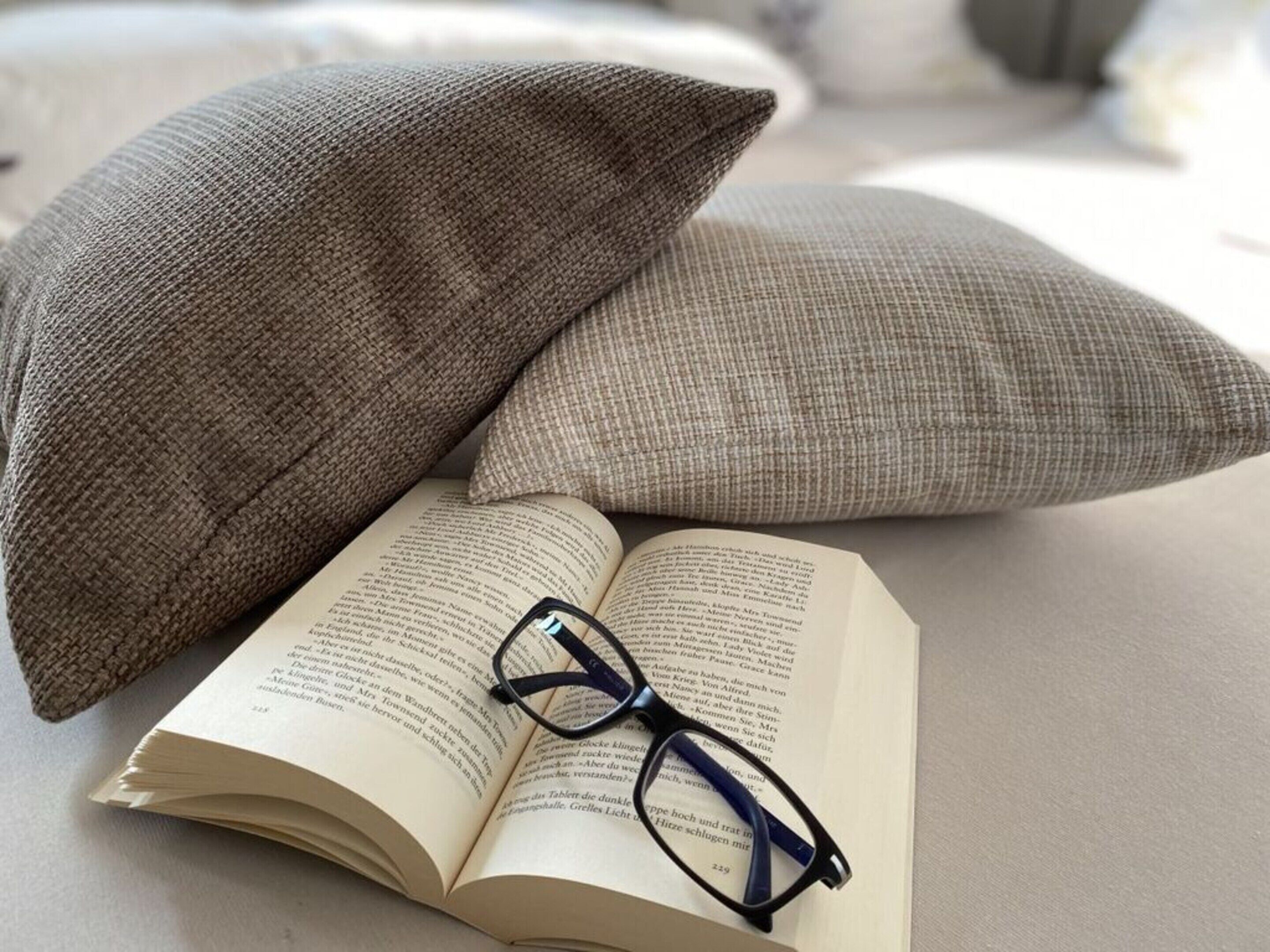 Un libro abierto con gafas encima, sobre una cama con dos almohadas.