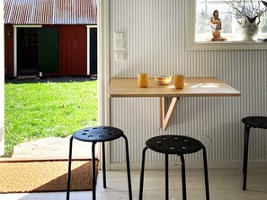 A room with a wooden table, two black stools, a window with a view of a garden, and a small statue on the windowsill.