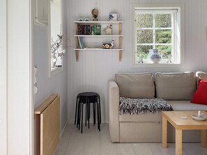 A room with a beige sofa, a wooden table, black stools, and a window with a view of greenery. Shelves with books and decorative items are mounted on the wall.
