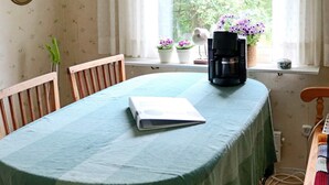 Un coin repas avec une table recouverte d’une nappe verte, une machine à café et des plantes en pot sur le rebord de la fenêtre.