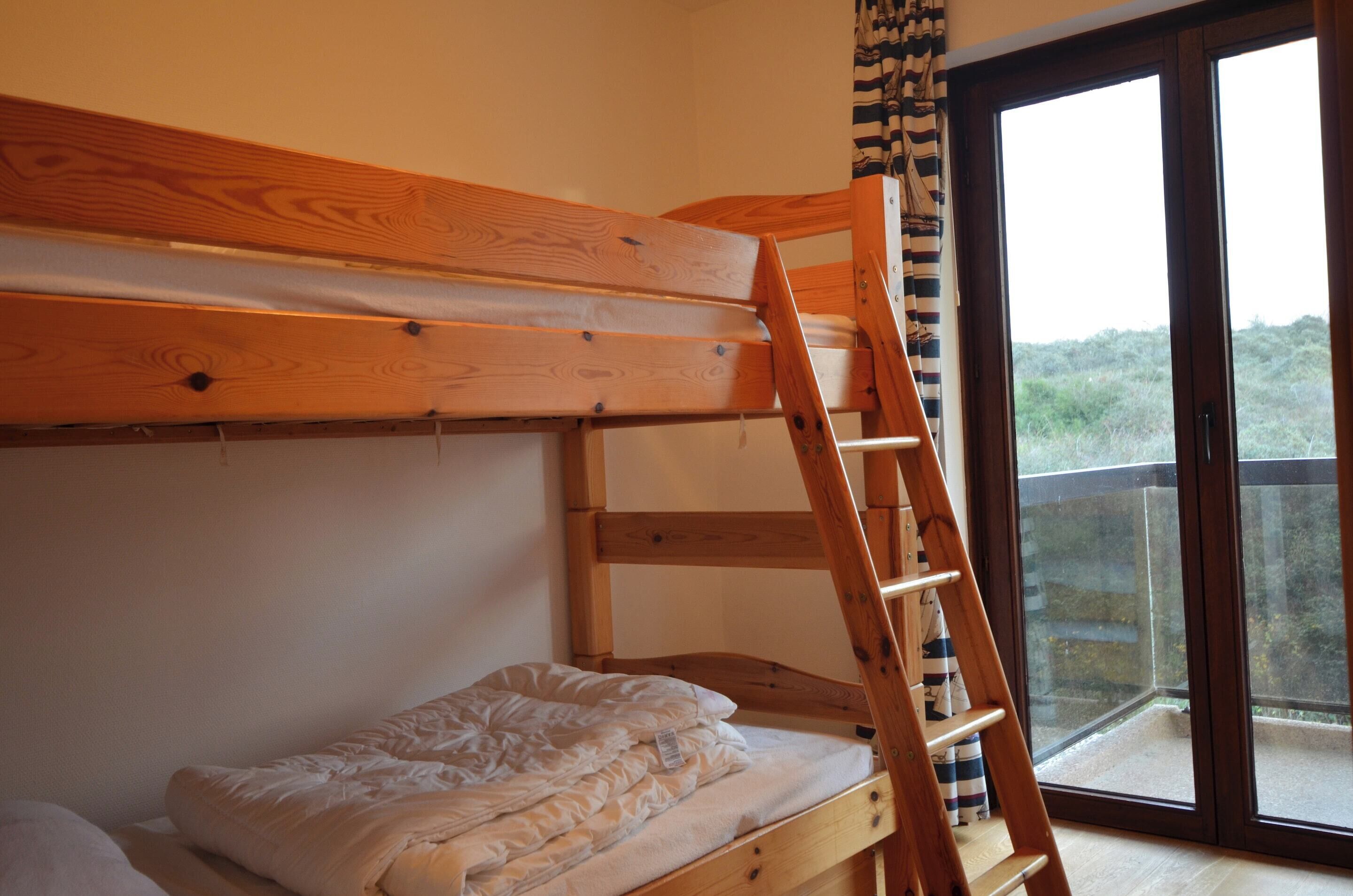 Un lit superposé avec une échelle en bois, un lit simple en dessous et une porte-fenêtre coulissante donnant sur un balcon extérieur.