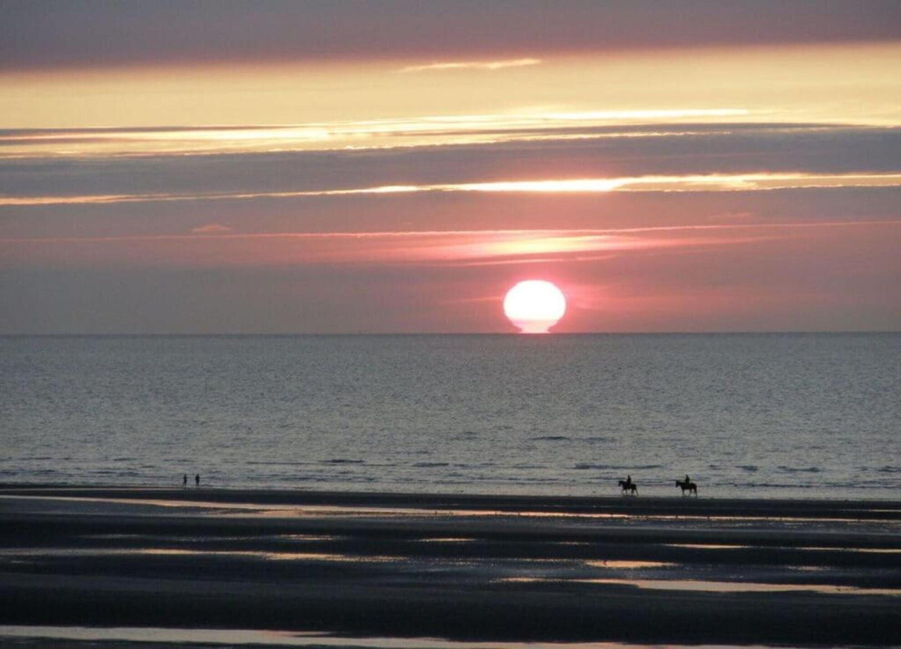 Un coucher de soleil sur une mer calme, avec quelques personnes et un cheval sur la plage.