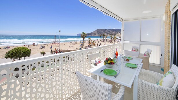 Un balcone con tavolo bianco apparecchiato per consumare un pasto, con vista su una spiaggia e palme.