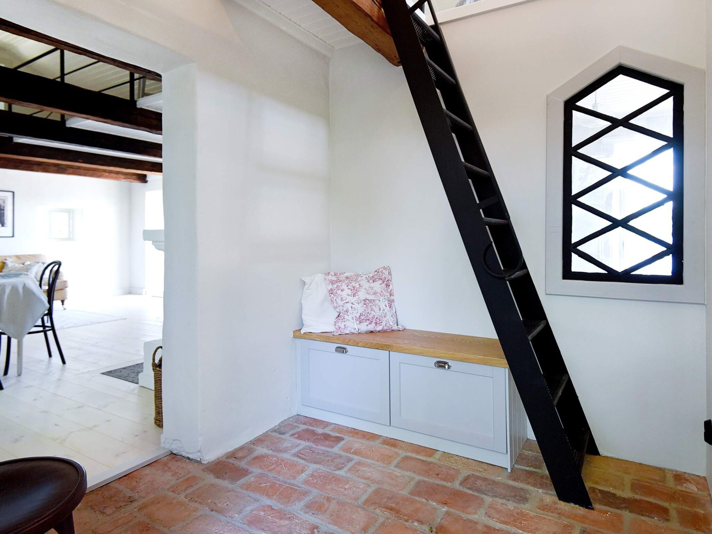 Um quarto com teto inclinado, um banco de madeira com almofada, um armário branco e uma janela com padrão geométrico.