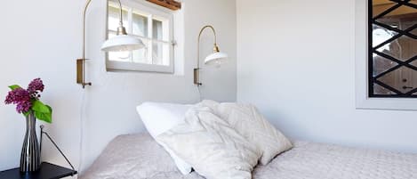 A bedroom with a bed, a window with a wooden beam, and two wall-mounted lamps.